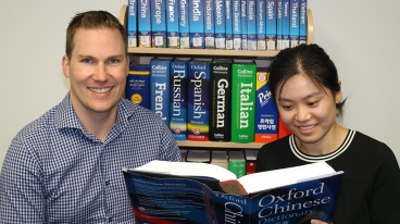 Language Faculty image - a man and a lady sitting in front of a bookshelf full of books about various countries, holding the Oxford Chinese Dictionary